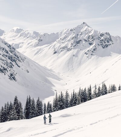 Skifahren in Gargellen | © Gargellner Bergbahnen, Daniel Zangerl Zwei Skifaher ziehen ihre Spuren durch eine verschneite Winterlandschaft in Gargellen. Sie bewegen sich am Rand eines weiten, weißen Berghangs oberhalb eines Nadelwaldes. Im Hintergrund erstreckt sich das imposante Valzifenztal, eingerahmt von steil aufragenden, schneebedeckten Gipfeln. Der Himmel ist klar und hell, durchzogen von einem Kondensstreifen – ein ruhiger, kraftvoller Moment inmitten der Natur. | © Gargellner Bergbahnen, Daniel Zangerl