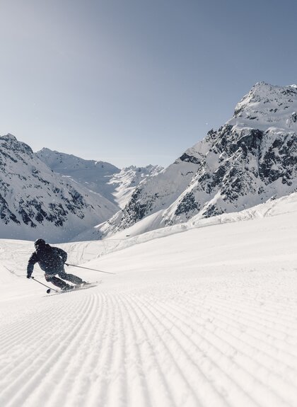 Skifahrer fährt dynamisch eine frisch präparierte Piste in Gargellen hinunter, Schneespritzer in der Luft, Berge im Hintergrund, die Madrisa und die Ritzenspitzen. | © Gargellner Berbhahnen GmbH & Co KG, Daniel Zangerl