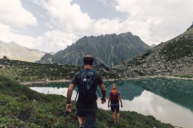 Zwei Wandernde nähern sich dem klaren Gandasee im Vergaldental. Das türkisfarbene Wasser spiegelt die umliegenden Felsen und die markanten Berggipfel wider, während Wolken den Himmel sanft bedecken – ein Moment ruhiger Bergidylle in den Montafoner Bergwelt. | © Gargellner Bergbahnen GmbH & Co KG, Daniel Zangerl