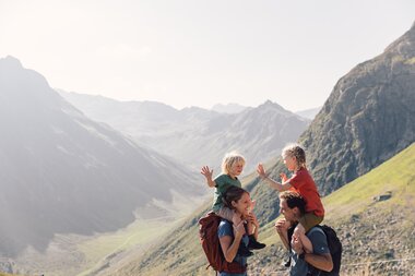 Eine Familie wandert am Gargellner Schafberg. Zwei Erwachsene tragen ihre Kinder auf den Schultern, während die Kinder sich lachend abklatschen. Im Hintergrund sind grüne Berghänge und ein tiefes Tal zu sehen. | © Gargellner Bergbahnen GmbH & Co KG, Daniel Zangerl