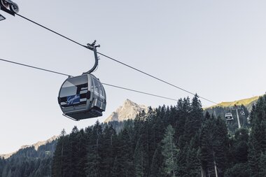 ine silberne Gondel der Gargellner Bergbahnen schwebt über einen dichten Nadelwald in Richtung Bergstation. Im Hintergrund ragt der Gipfel der Madrisa in der Abendsonne auf, während der Himmel klar und hellblau ist. | © Gargellner Bergbahnen GmbH & Co KG, Daniel Zangerl