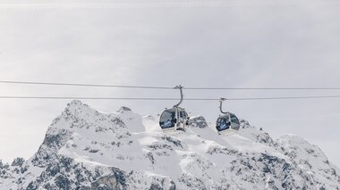 Zwei Gondeln der Schafbergbahn in Gargellen schweben vor einer beeindruckenden, schneebedeckten Bergkulisse. Die modernen Kabinen der Gargellner Bergbahnen heben sich klar vom hellen Winterhimmel ab, während im Hintergrund die majestätischen Gipfel der Montafoner Alpen aufragen. | © Gargellner Bergbahnen GmbH & Co KG, Daniel Zangerl