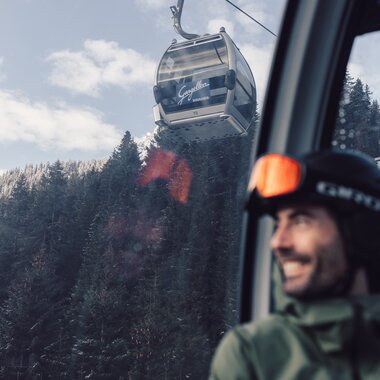 Ein Skifahrer mit Helm und Skibrille sitzt in einer Gondel der Schafbergbahn in Gargellen. Durch das Fenster ist die Seilbahn zu sehen, die über verschneite Wälder und Hänge führt. Im Hintergrund steigen die Gondeln der Gargellner Bergbahnen den schneebedeckten Berg hinauf. | © Gargellner Bergbahnen GmbH & Co KG, Daniel Zangerl