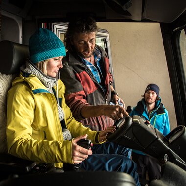 ine Frau in gelber Jacke sitzt in der Fahrerkabine einer Pistenraupe, während ein Mitarbeiter der Gargellner Bergbahnen ihr die Steuerung erklärt. Ein weiterer Kollege beobachtet das Gespräch interessiert. Die Szene zeigt den Einblick hinter die Kulissen des Skigebiets und die technische Schulung an den Maschinen. | © Gargellner Bergbahnen GmbH & Co KG, Christoph Schöch
