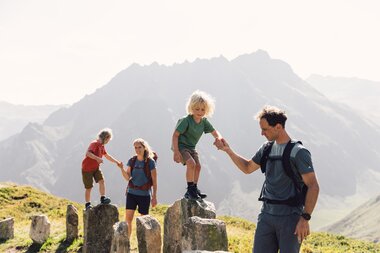 ine Familie wandert entlang großer Felsen und balanciert spielerisch über die Steine. Die Kinder werden von den Eltern an der Hand gehalten, während im Hintergrund die Berge des Montafon leuchten. | © Gargellner Bergbahnen GmbH & Co KG, Daniel Zangerl