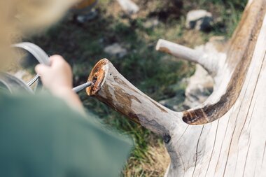 Detailaufnahme eines hölzernen Wasserlaufs in den Bergen von Gargellen. Ein Kind gießt Wasser aus einer Gießkanne in die Rinne, das langsam durch den geschnitzten Baumstamm fließt. Das helle Holz und die Wassertropfen schimmern in der Sonne. | © Gargellner Bergbahnen GmbH & Co KG, Daniel Zangerl