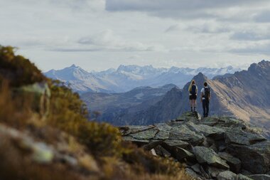 Zwei Wandernde stehen auf einem Felsen hoch über Gargellen und genießen den Blick auf die umliegenden Berge. Der Herbst taucht die Landschaft in sanfte Farben, während die schneebedeckten Gipfel in der Ferne leuchten – ein Moment der Ruhe und Weite in Gargellen. | © Gargellner Bergbahnen GmbH & Co KG, Alexander Furchs 