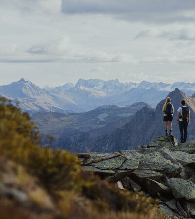 Weitblick über die Montafoner Bergwelt | © Gargellner Bergbahnen GmbH & Co KG, Alexander Furchs Zwei Wandernde stehen auf einem Felsen hoch über Gargellen und genießen den Blick auf die umliegenden Berge. Der Herbst taucht die Landschaft in sanfte Farben, während die schneebedeckten Gipfel in der Ferne leuchten – ein Moment der Ruhe und Weite in Gargellen. | © Gargellner Bergbahnen GmbH & Co KG, Alexander Furchs
