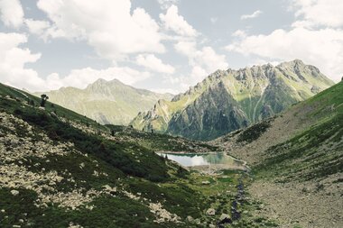 Der Gandasee liegt ruhig eingebettet zwischen steilen Felswänden und grünen Hängen oberhalb von Gargellen. Im klaren Wasser spiegeln sich Wolken und Gipfel, während ein kleiner Bach aus dem Tal heraufplätschert. Eine eindrucksvolle Szenerie, die die alpine Ursprünglichkeit des Montafons widerspiegelt. | © Gargellner Bergbahnen GmbH & Co KG, Daniel Zangerl