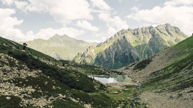 Der Gandasee | © Gargellner Bergbahnen GmbH & Co KG, Daniel Zangerl Der Gandasee liegt ruhig eingebettet zwischen steilen Felswänden und grünen Hängen oberhalb von Gargellen. Im klaren Wasser spiegeln sich Wolken und Gipfel, während ein kleiner Bach aus dem Tal heraufplätschert. Eine eindrucksvolle Szenerie, die die alpine Ursprünglichkeit des Montafons widerspiegelt. | © Gargellner Bergbahnen GmbH & Co KG, Daniel Zangerl
