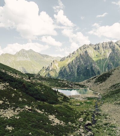 Der Gandasee | © Gargellner Bergbahnen GmbH & Co KG, Daniel Zangerl Der Gandasee liegt ruhig eingebettet zwischen steilen Felswänden und grünen Hängen oberhalb von Gargellen. Im klaren Wasser spiegeln sich Wolken und Gipfel, während ein kleiner Bach aus dem Tal heraufplätschert. Eine eindrucksvolle Szenerie, die die alpine Ursprünglichkeit des Montafons widerspiegelt. | © Gargellner Bergbahnen GmbH & Co KG, Daniel Zangerl