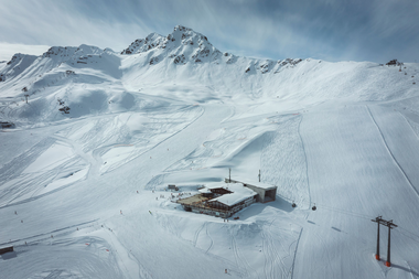 Luftaufnahme des Schafbergplateaus in Gargellen mit verschneiten Skipisten, Skifahrern und der Bergstation der Gondelbahn im Vordergrund. Im Hintergrund erheben sich schneebedeckte Gipfel unter leicht bewölktem Himmel. | © Gargellner Bergbahnen GmbH & Co KG 