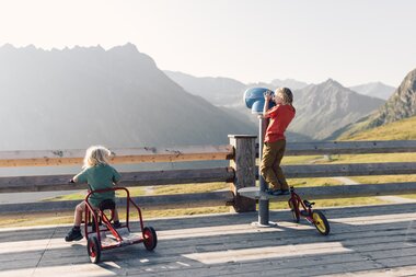 Zwei Kinder spielen auf einer Holzterrasse mit Blick auf die Berge von Gargellen. Ein Kind sitzt auf einem roten Dreirad und schaut über das Geländer, während das andere Kind auf einem kleinen Tritt steht und durch ein fest installiertes blaues Fernrohr blickt. Im Hintergrund sind grüne Berghänge und die Silhouetten der Gipfel in der Abendsonne zu sehen. | © Gargellner Bergbahnen GmbH & Co KG, Daniel Zangerl
