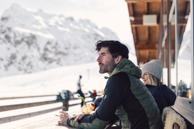 Zeit für eine Pause im Schafberg Hüsli | © Gargellner Bergbahnen GmbH & Co KG, Daniel Zangerl Eine Person in grüner Winterjacke sitzt an einem Holztisch auf einer Sonnenterrasse und blickt gedankenverloren in die Ferne. Neben ihr sitzt eine weitere Person mit grauer Mütze und dunkler Jacke. Im Hintergrund sind schneebedeckte Berge und ein Holzgebäude zu sehen. | © Gargellner Bergbahnen GmbH & Co KG, Daniel Zangerl