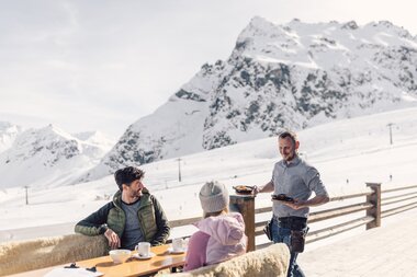 Mittagessen mit Bergblick auf der Sonnenterrasse des Schafberg Hüsli | © Gargellner Bergbahnen GmbH & Co KG, Daniel Zangerl Ein Kellner serviert zwei Gästen auf einer Sonnenterrasse im Skigebiet das Essen. Die Gäste sitzen an einem Holztisch mit Kaffee und genießen den Blick auf die schneebedeckten Berge im Hintergrund. Der Himmel ist klar und die Sonne beleuchtet die Winterlandschaft. | © Gargellner Bergbahnen GmbH & Co KG, Daniel Zangerl