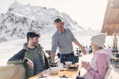 Ein Kellner serviert zwei Gästen auf einer sonnigen Terrasse im Skigebiet das Mittagessen. Die Gäste sitzen an einem Holztisch mit warmen Getränken und lächeln. Im Hintergrund sind schneebedeckte Berge und ein klarer Winterhimmel zu sehen. | © Gargellner Bergbahnen GmbH & Co KG, Daniel Zangerl