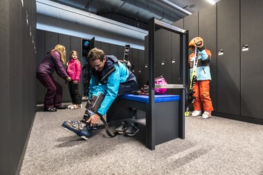 Skidepot direkt bei der Talstation der Gargellner Bergbahnen | © Gargellner Bergbahnen GmbH & Co KG, Christoph Schöch Eine Familie bereitet sich im Skidepot auf den Skitag vor. Ein Mann sitzt auf einer Bank und zieht seine Skischuhe an, während ein Kind im orange-blauen Skianzug mit Helm und Skiern daneben steht. Im Hintergrund helfen eine Frau und ein weiteres Kind beim Anziehen der Ausrüstung. Umgeben sind sie von grauen Spinden und Sitzgelegenheiten. | © Gargellner Bergbahnen GmbH & Co KG, Christoph Schöch
