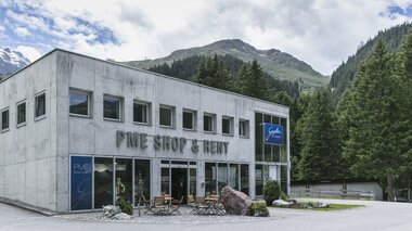 Das Gebäude des PME Shop & Rent in Gargellen steht vor einer Kulisse aus grünen Bergen und dichten Wäldern. Das zweistöckige Haus aus grauem Beton hat große Glasfronten im Erdgeschoss mit der Aufschrift „PME Shop & Rent“ sowie dem blauen Schild „Gargellen Montafon“. Vor dem Eingang stehen Tische und Stühle auf einer kleinen Terrasse. | © Gargellner Bergbahnen GmbH & Co KG, Christoph Schöch