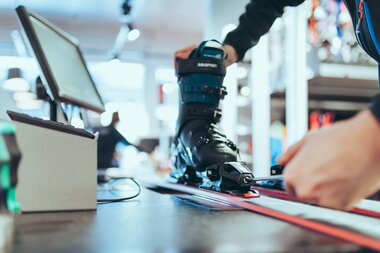 Bindungseinstellung im PME Shop & Rent | © Gargellner Bergbahnen GmbH & Co KG, Christoph Schöch Eine Person stellt mit einem Schraubenzieher die Bindung eines Skis ein, in dem ein schwarzer Skischuh der Marke Salomon eingespannt ist. Der Ski liegt auf einer Werkbank in einem modernen Skiverleih. Im Hintergrund sind Arbeitsgeräte und Regale leicht unscharf zu erkennen. | © Gargellner Bergbahnen GmbH & Co KG, Christoph Schöch
