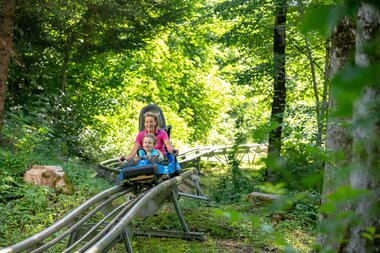 Alpine-Coaster-Golm im Montafon | © Golm Silvretta Lünersee Tourismus GmbH Bregenz, Stefan Kothner Eine Frau und ein Kind fahren auf einer Sommerrodelbahn durch den Wald hinunter | © Golm Silvretta Lünersee Tourismus GmbH Bregenz, Stefan Kothner